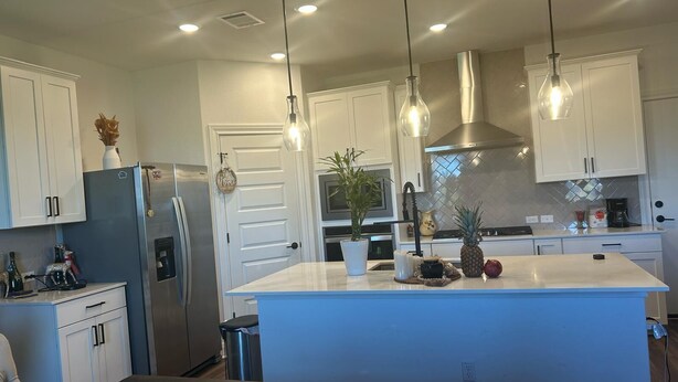 A modern kitchen with white cabinets, a stainless steel refrigerator, a blue island, and pendant lighting. - New house. 20 minutes from ATX, 5 bedrooms (Manor)