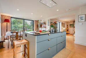 A modern kitchen with a central island, wooden dining table, and chairs. There is a large window providing a view of greenery outside. - Spacious cottage overlooking the fields (Milford on Sea)
