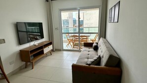 A living room with a flat-screen TV, a wooden TV stand, a beige sofa, and a sliding glass door leading to an outdoor balcony with wooden furniture. - Apartment in Ubatuba Tenório. 3 minutes from the beach. (Ubatuba)