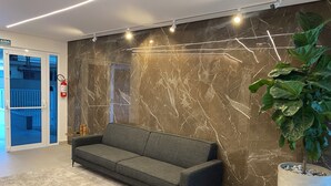 A modern lobby with a marble wall, a grey sofa, a potted plant, and a fire extinguisher. - Apartment in Ubatuba Tenório. 3 minutes from the beach. (Ubatuba)