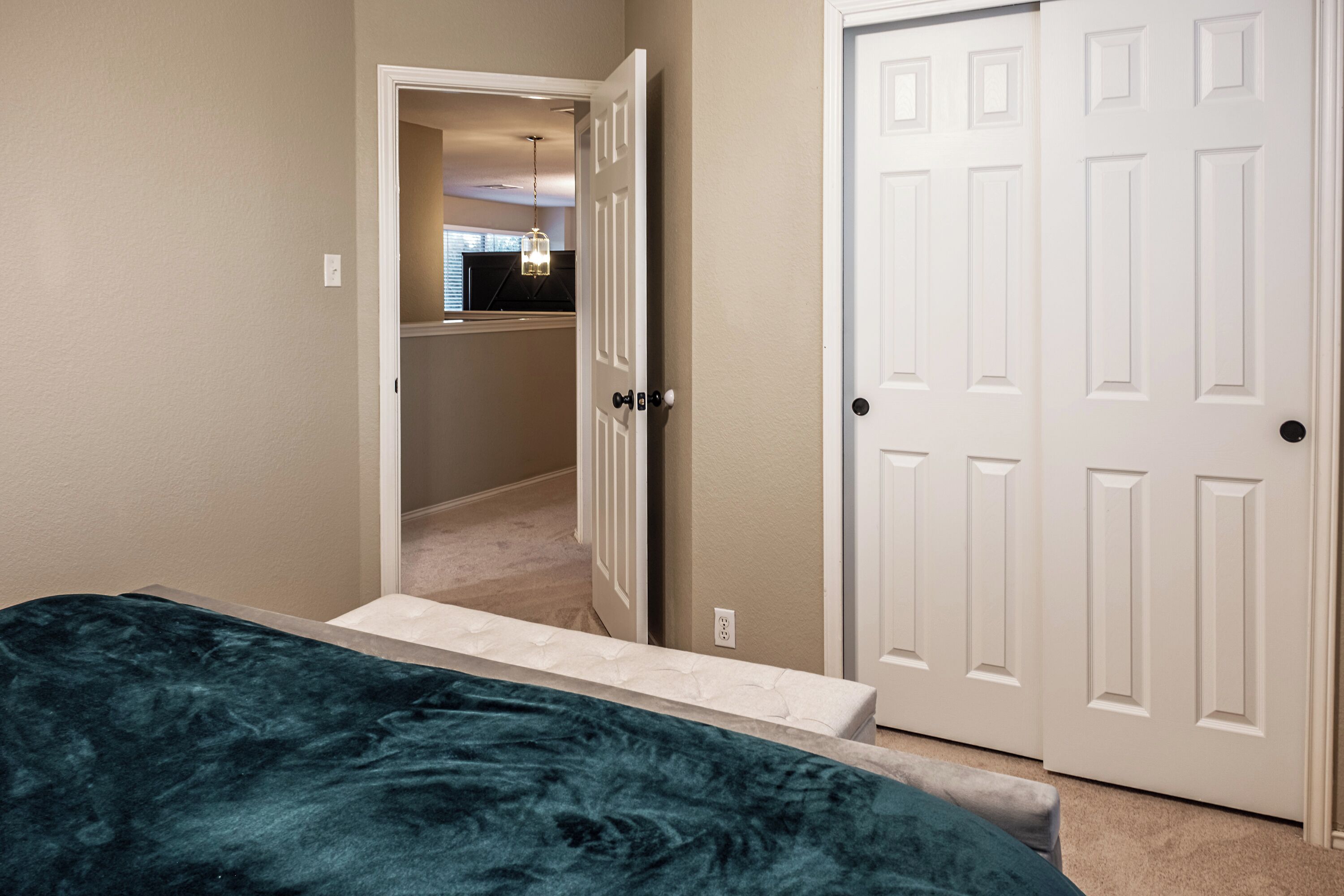 A bedroom with a bed, a white door, and a hallway leading to another room.