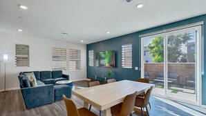 Una sala de estar moderna con un sofá, una mesa de comedor y sillas. Hay un ventanal con persianas, un cuadro en la pared y una rejilla de ventilación en el techo.