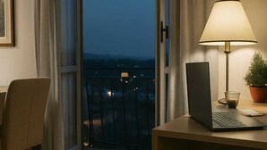 Una habitación con una computadora portátil sobre un escritorio de madera, una lámpara, una silla, una planta en maceta y vistas a través de un amplio ventanal.