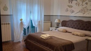 Un dormitorio con cama, mesita de noche, lámpara, radiador y ventana con cortinas translúcidas.