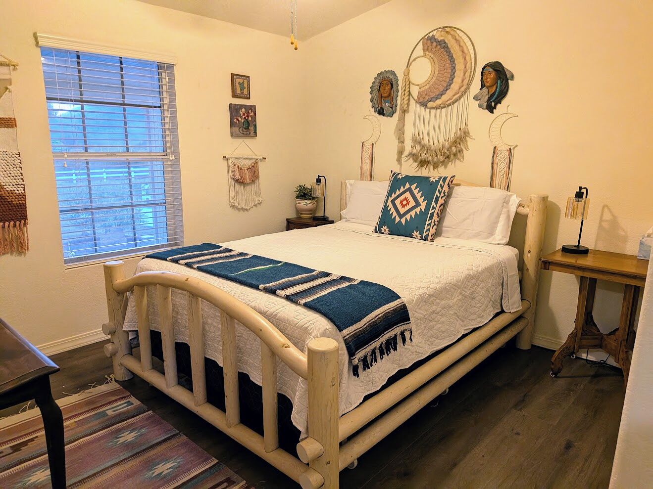 Un dormitorio con cama de madera, una ventana con persianas, una mesita de noche y adornos en la pared, incluyendo atrapasueños y una diadema con atrapasueños.