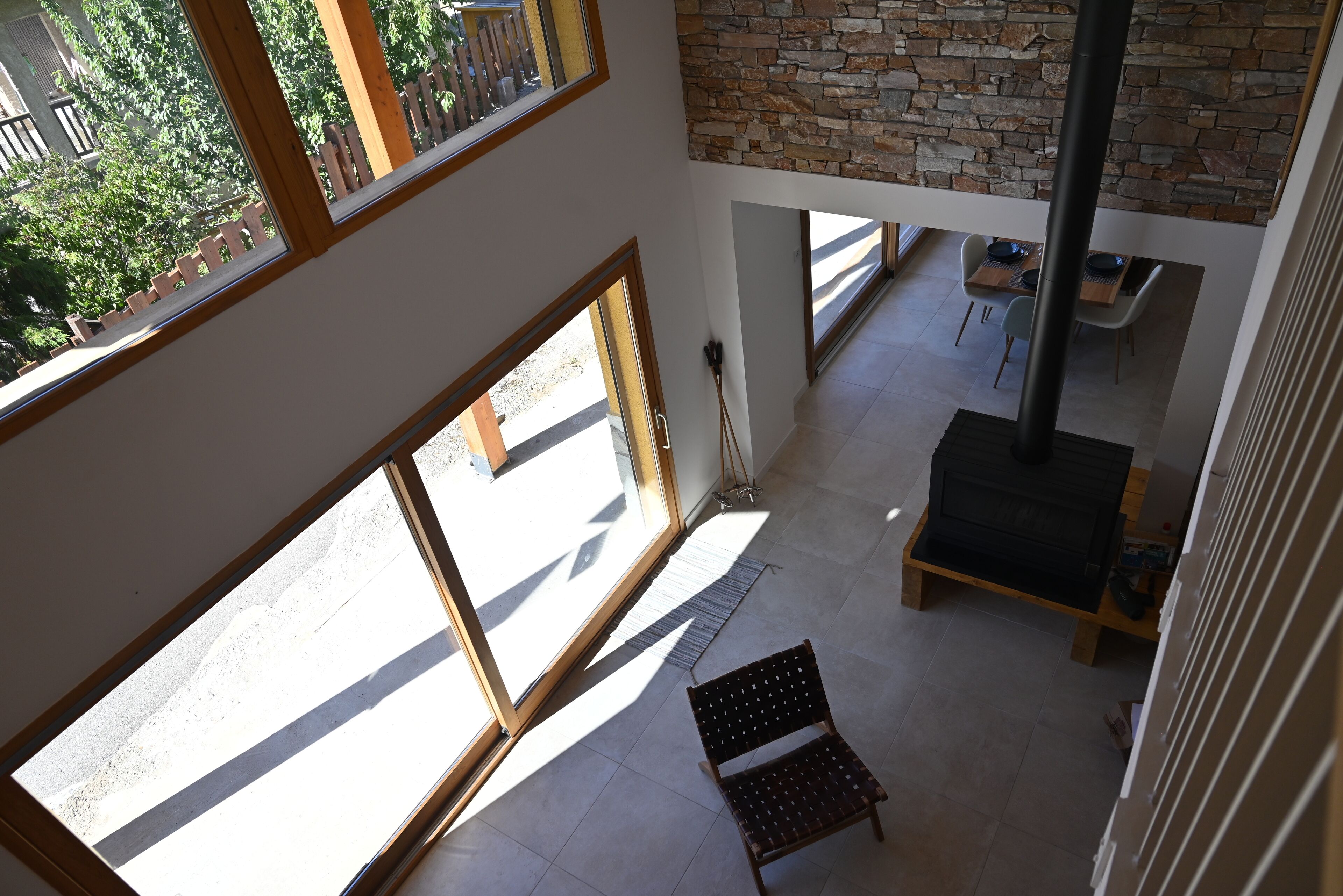 A high-ceilinged room with large windows, a stone wall, a fireplace, and a dining area.