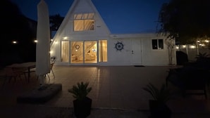Ein weißes, A-Haus mit der auf der Tür deutlich sichtbaren Nummer '451', einem Schiffswellenrad an der Wand und einer Treppe, die zum oberen Stockwerk führt.