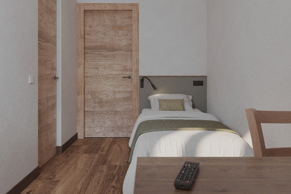 A small, neatly arranged bedroom with a wooden door, a single bed with a headboard, a bedside table with a remote control, and a wooden floor.
