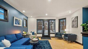 Ein modernes Wohnzimmer mit einem blauen Sofa, einem Couchtisch und einem blauen Teppich. Zwei Türen führen auf einen Balkon, und ein kleiner Tisch mit Lampe und Pflanze steht dort.