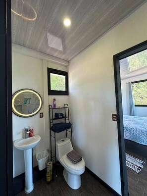 Ein kleines Badezimmer mit Toilette, Waschbecken und einem Spiegel mit Beleuchtung. Über der Toilette befindet sich ein Fenster und eine Tür, die auf einen Balkon führt.