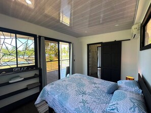 Ein Schlafzimmer mit einem Bett, einem Fenster mit Blick auf Palmen und einer Tür zu einem anderen Raum.