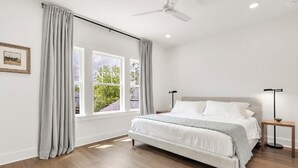 Et soveværelse med seng, ventilator, natbord og et vindue med gardiner.