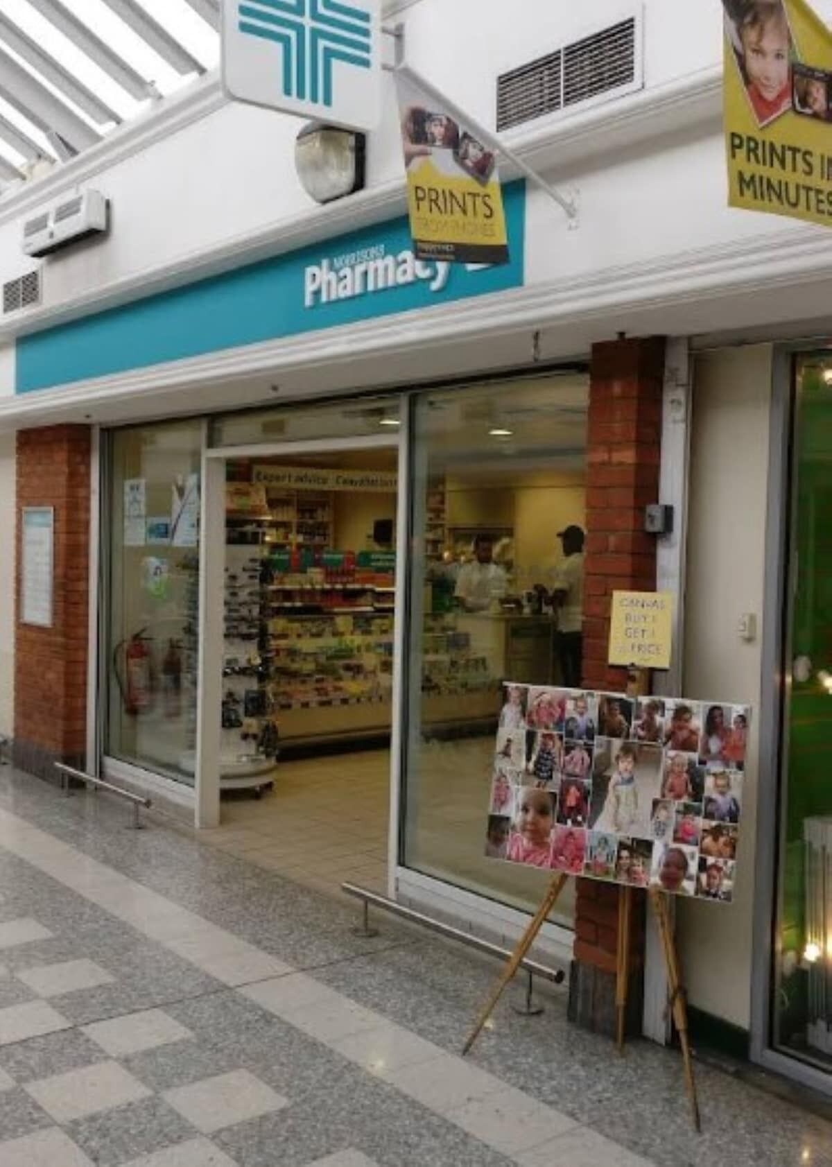 A pharmacy with a blue sign, a display of photos on a stand, and a sign indicating a buy-one-get-one-free offer.