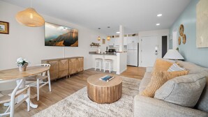 Ein modernes Wohnzimmer mit einem Holzesstisch, weißen Stühlen, einem runden Couchtisch und einer Couch mit Kissen. Im Hintergrund ist eine Küche mit weißen Schränken und einer Mikrowelle erkennbar.