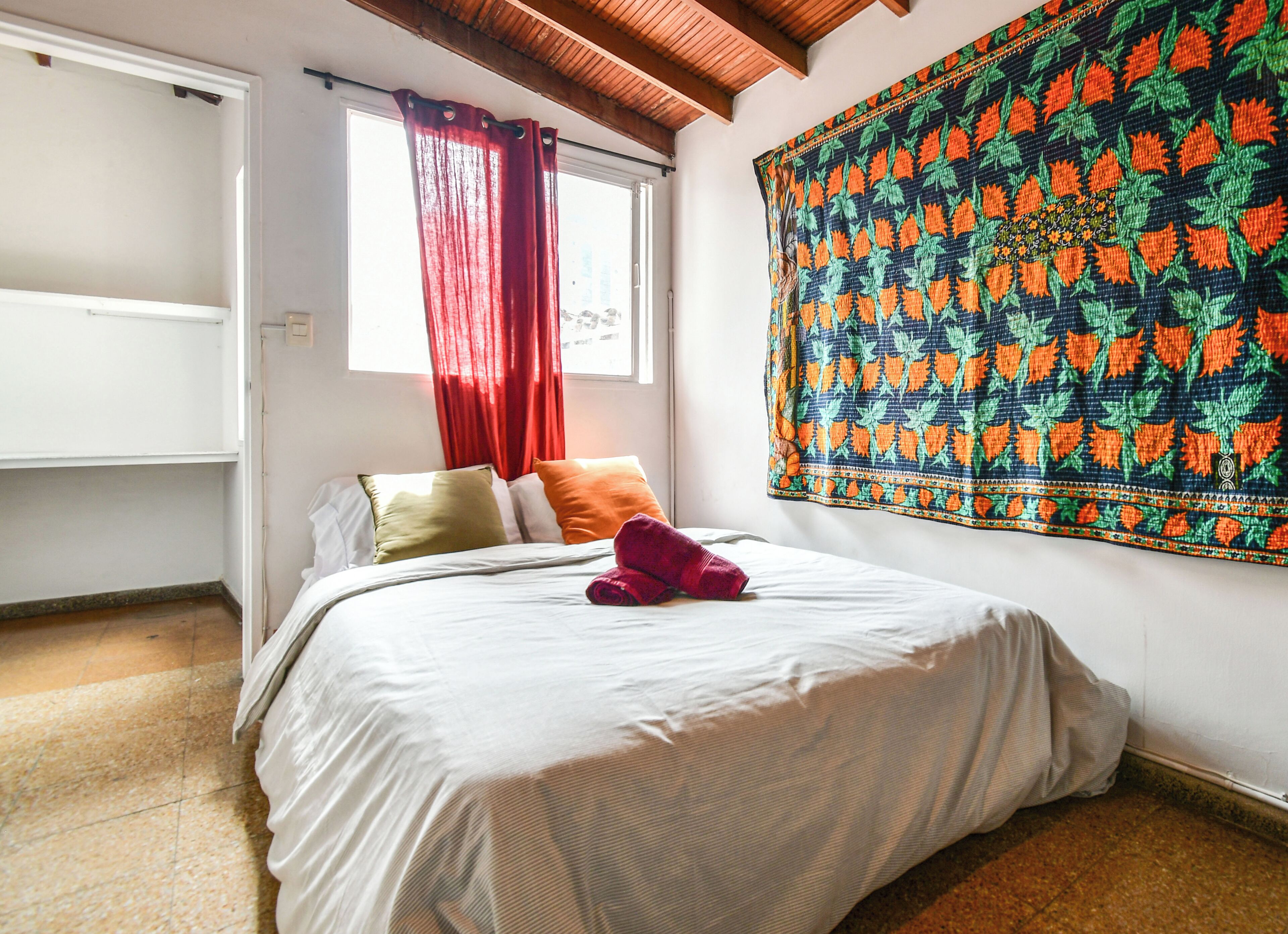 A bedroom with a bed, pillows, a red curtain, a tapestry, and a window.