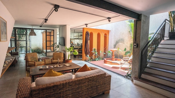 A spacious living area with a large wicker sofa, a coffee table, and a dining table. There is a kitchen visible in the background, and a staircase leading to an outdoor area with plants. - Casa Bolivar by Wynwood House (Medellín)