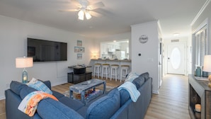 Ein modernes Wohnzimmer mit grauem Sofa, einem Couchtisch und einem an der Wand montierten Fernseher. Im Hintergrund befindet sich ein Küchenbereich mit weißen Schränken und einem Barhocker.