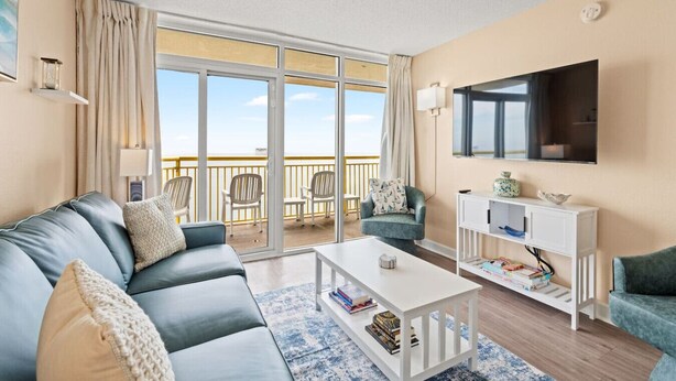 Un salon moderne avec un canapé, des fauteuils, une table basse et un téléviseur. Il y a un balcon avec vue sur le ciel.