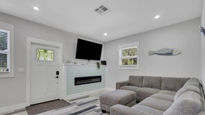 Een moderne woonkamer met een grijze bank, een flatscreen televisie aan de muur, een open haard met een lichtblauwe schoorsteenmantel en een vloer met een patroon.