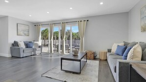 A spacious living room with a grey sofa, a coffee table, and a large sliding glass door leading to an outdoor patio. - Spacious 4BR Home w/ Heated Pool & Pickleball Court on 5-Acre Estate (Naples)