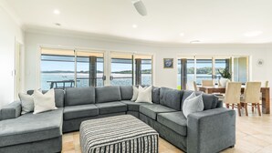 Un salon moderne avec un canapé d’angle gris, un pouf à rayures et un coin repas comprenant des chaises et une table en bois. De grandes fenêtres offrent une vue sur l’eau.