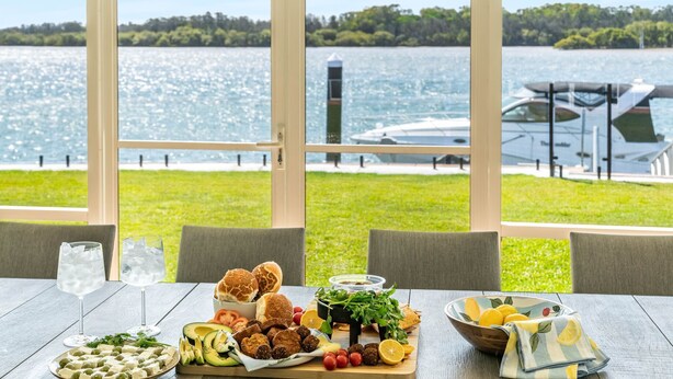 Une table dressée avec une variété de plats et de boissons, donnant sur un lac où l’on peut voir des bateaux et une végétation luxuriante.