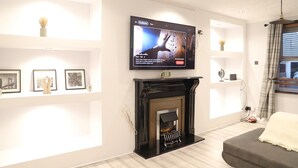 A modern living room with a fireplace, a flat-screen TV mounted above it, and framed pictures on the walls. - Argyleluxe (Worsley)