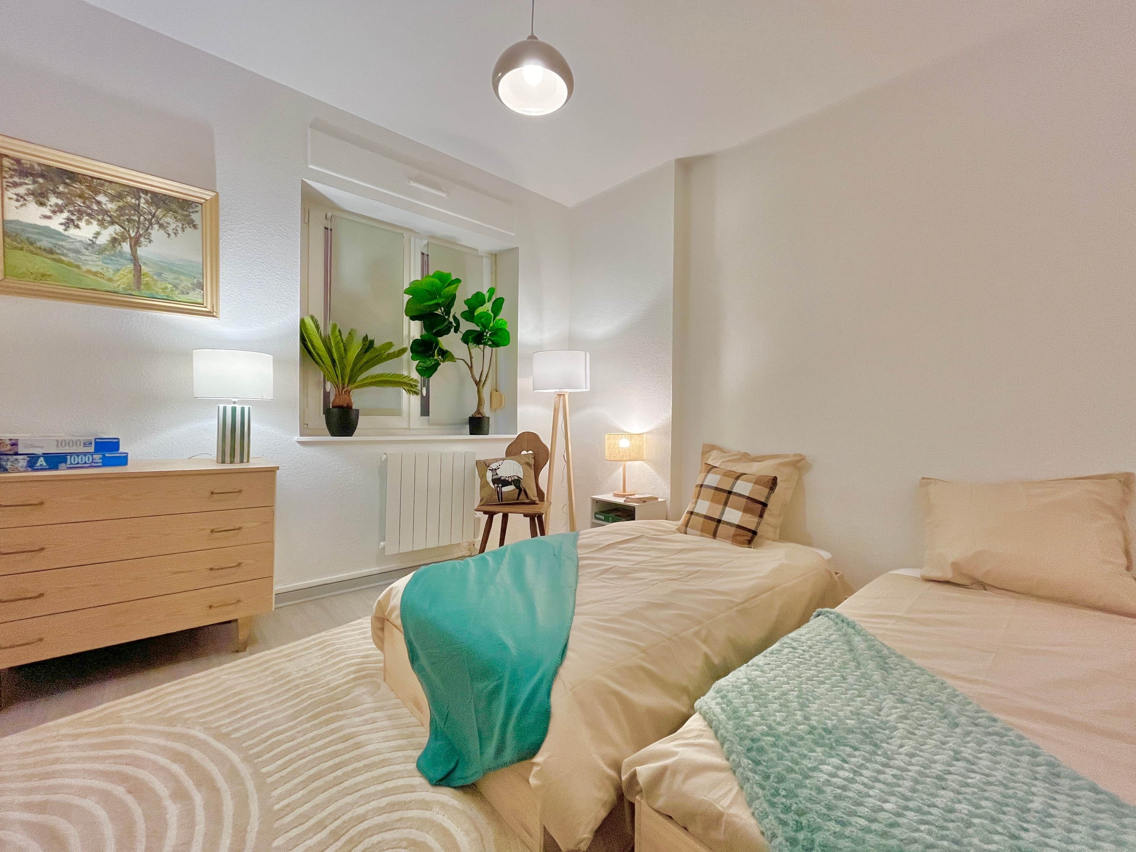 Une chambre à coucher avec un lit, une commode, un miroir, deux plantes en pot, une lampe et une fenêtre.