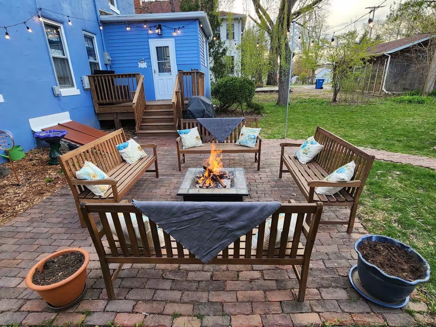Une terrasse avec des meubles en bois, un foyer extérieur et une maison bleue en arrière-plan.