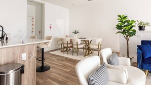 Un salón moderno con comedor, que presenta suelo de madera, mobiliario blanco y una planta en maceta.