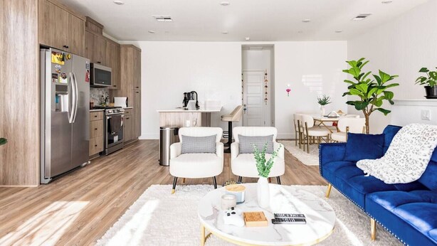 Una sala moderna con un sofá azul, sillones blancos y una mesa de centro. Hay una zona de cocina con un refrigerador y una mesa de comedor.