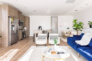 A modern living room with a blue sofa, white armchairs, and a coffee table. There is a kitchen area with a refrigerator and a dining table. - Clovis Retreat - Modern 2BR Near Parks, Pool (Clovis)