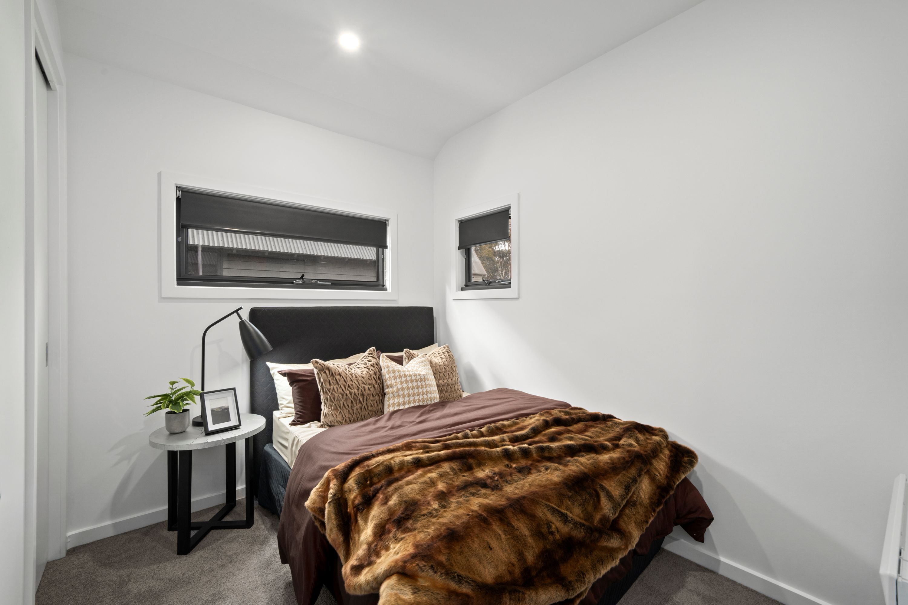 A bedroom with a bed, a small table with a plant, and two windows.