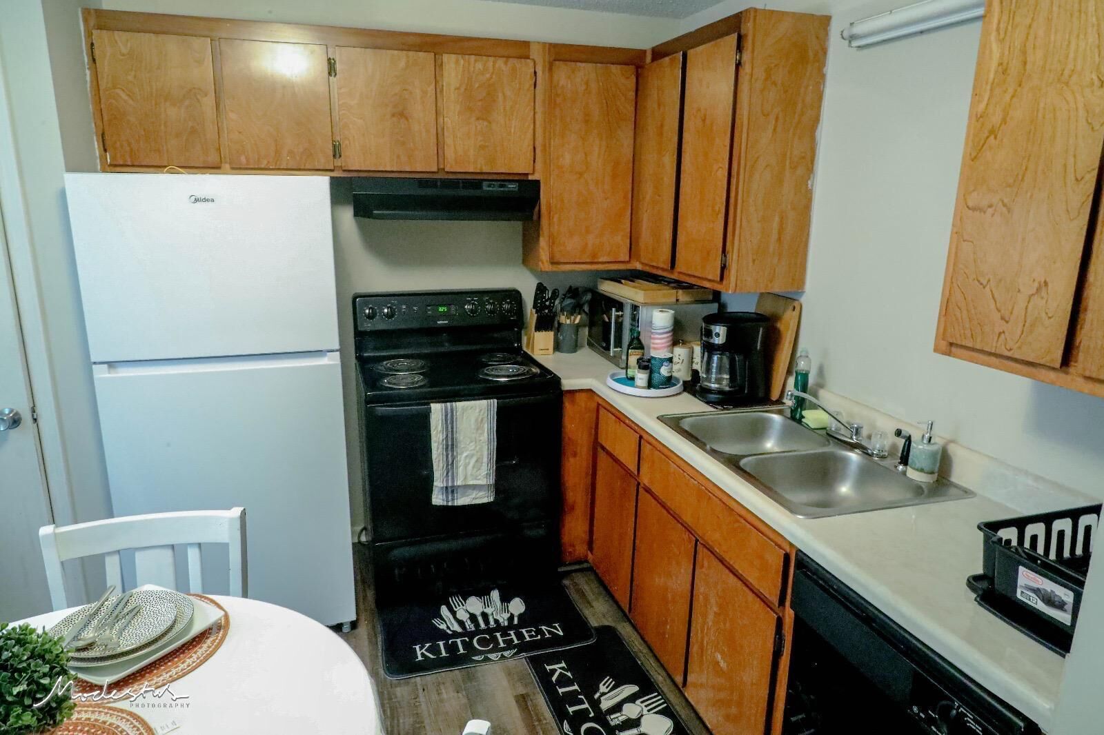 A compact kitchen with wooden cabinets, a black stove, a white refrigerator, and a stainless steel sink.