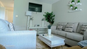 Ein modernes Wohnzimmer mit grauem Sofa, weißem Couchtisch und einem an der Wand montierten Fernseher. Weiße Regale mit Pflanzen und dekorativen Objekten sowie ein weißer Konsolentisch mit Büchern und einer Lampe.