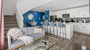 A modern living room with a gray sofa, a coffee table, and a kitchen area with white cabinets and a blue accent wall. - 1 King Loft W/Parking + Laundry I Oak Lawn + DT (Dallas)