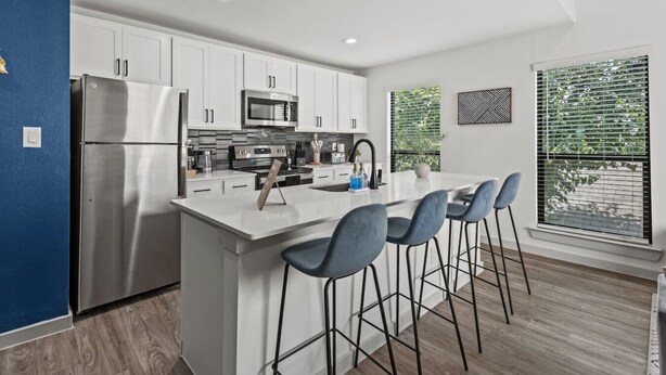 A modern kitchen with white cabinetry, stainless steel appliances, a central island with bar stools, and a large window with blinds. - 1 King Loft W/Parking + Laundry I Oak Lawn + DT (Dallas)
