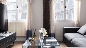 Ett modernt vardagsrum med ett soffbord i glas, en soffa, en tv och utsikt över byggnader genom stora fönster.