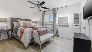 Een slaapkamer met een bed, nachtkastje, stoel, plafondventilator, raam met gordijnen, spiegel, televisie en een kledingkast.