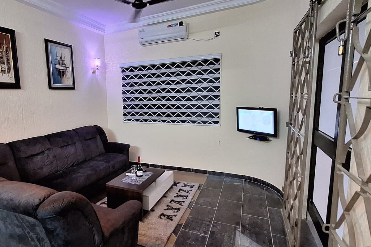 Een woonkamer met een donkerbruine bank, een klein bijzettafeltje en een vloerkleed met dessin. Er is een raam met metalen tralies, een wandairconditioner en een televisie op een standaard.