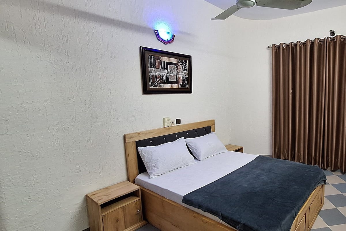 Een slaapkamer met een bed, nachtkastje, wandlamp, plafondventilator, raam met gordijnen en een lijst met een afbeelding aan de muur.