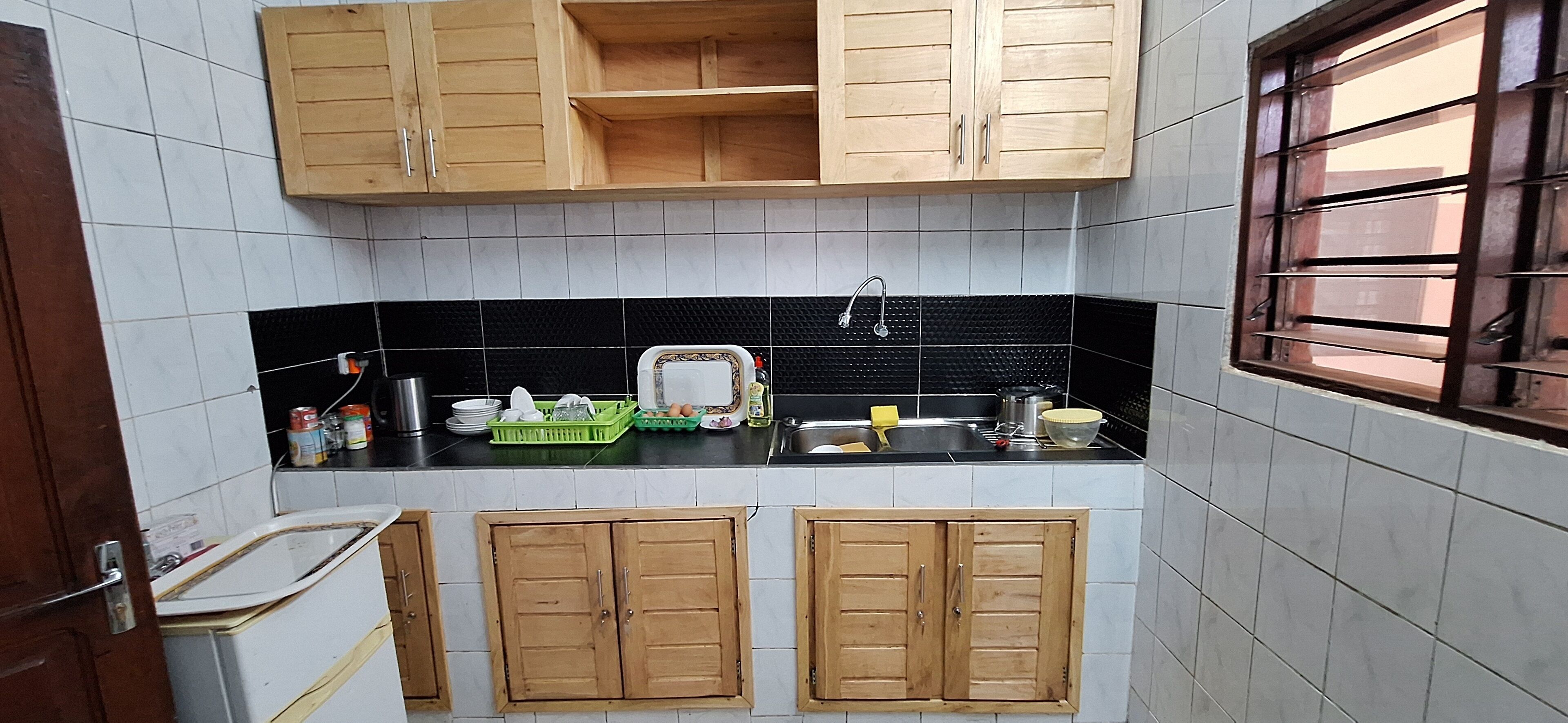 Een kleine keuken met houten kastjes, een zwart aanrecht en een raam met tralies.