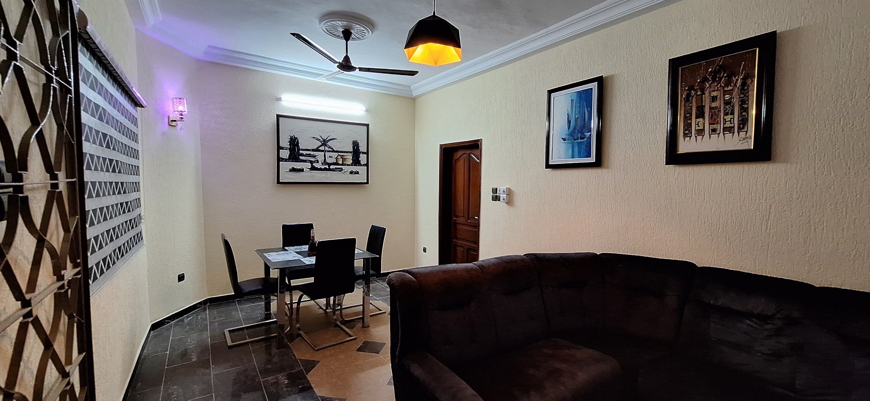 Una sala de estar con un sofá marrón, una mesa de comedor con sillas, un ventilador de techo y obras de arte enmarcadas en las paredes.