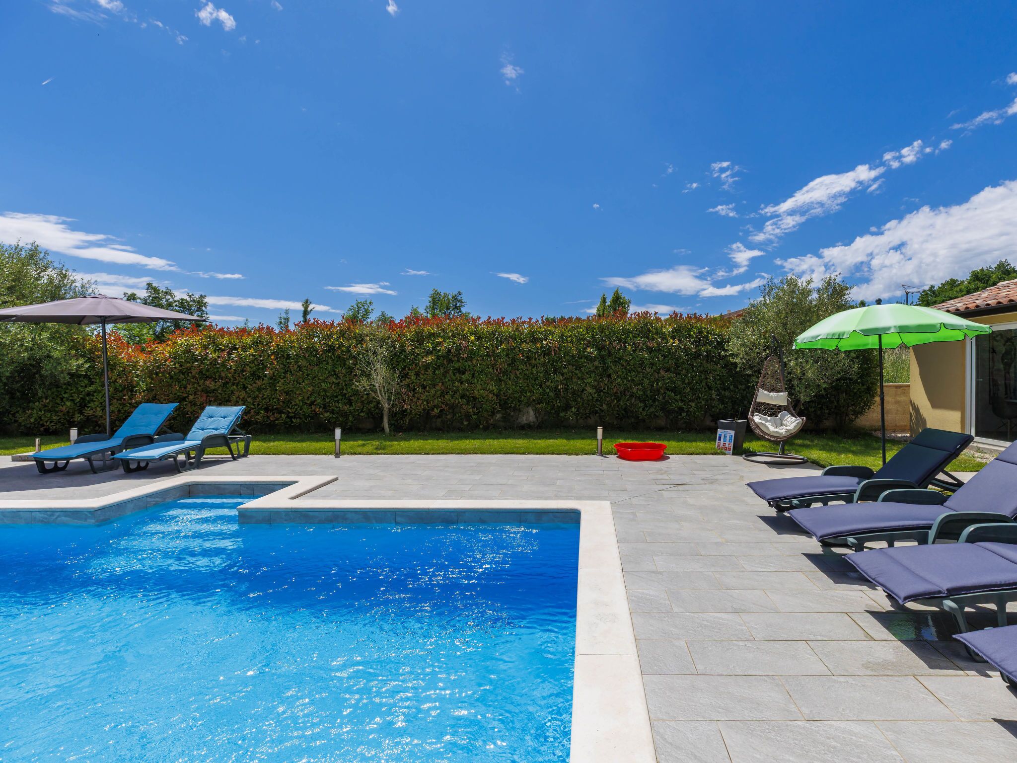 A swimming pool with lounge chairs and umbrellas, surrounded by a hedge and a tiled patio.