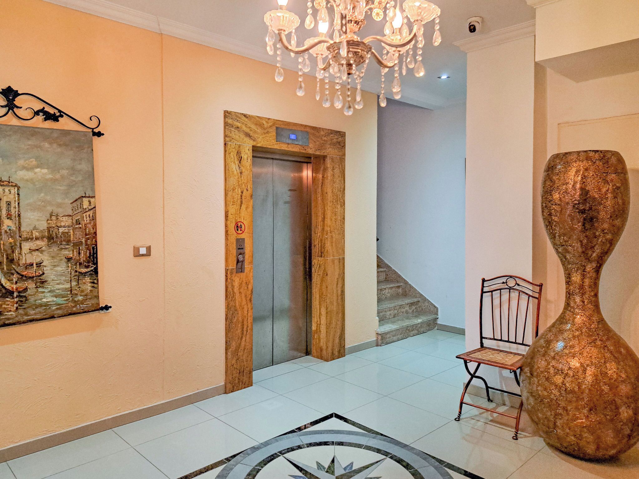 A hallway with a wooden elevator, a chandelier, a large vase, and a painting of a cityscape.