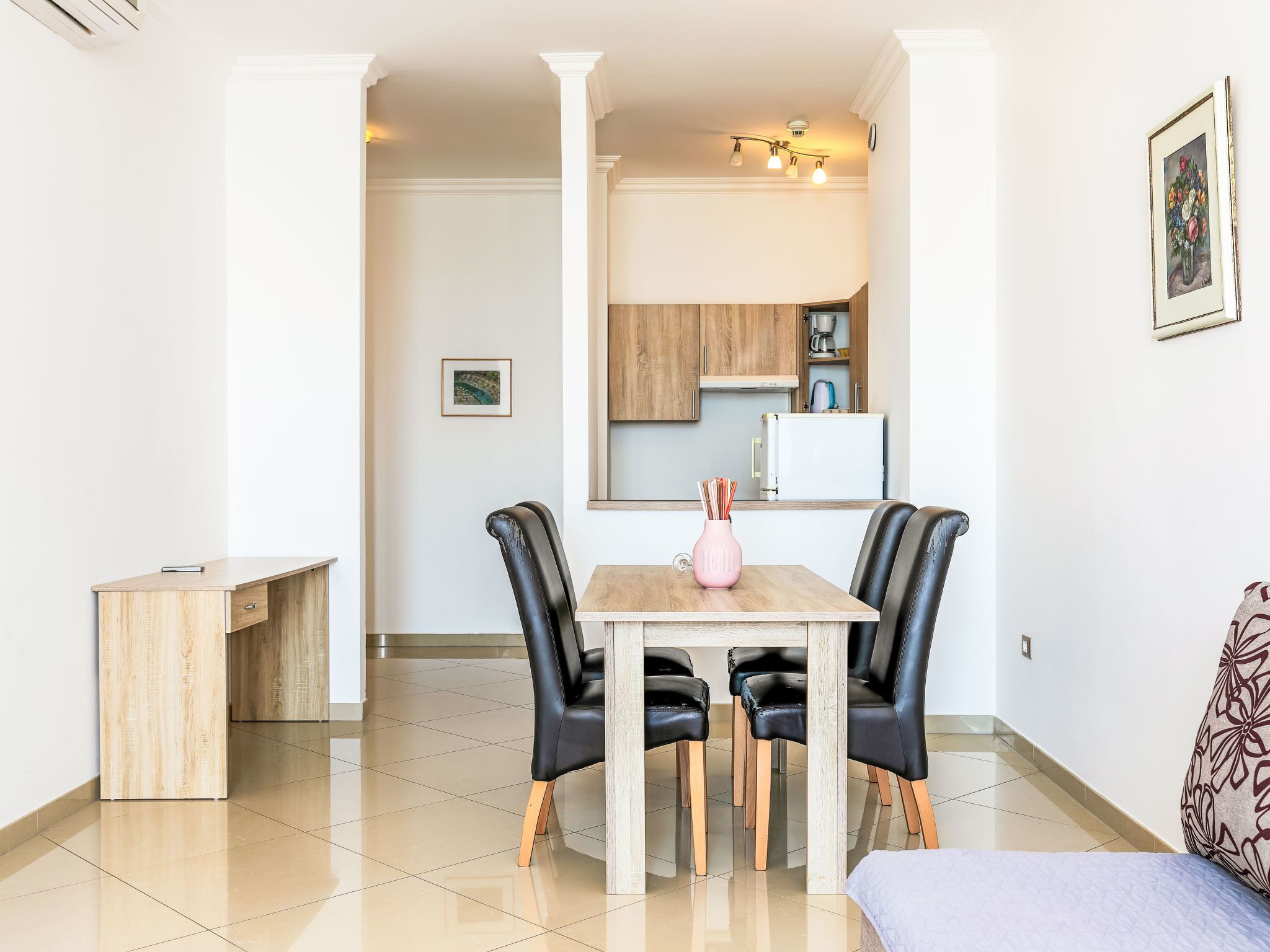 Un espace repas moderne avec une table en bois et des chaises noires, un petit vase rose et un tableau encadré accroché au mur.