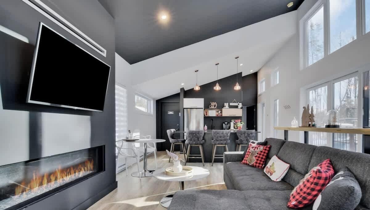 A modern living room with a flat-screen TV, fireplace, grey sofa with red and black pillows, dining area with chairs, and a kitchen in the background.