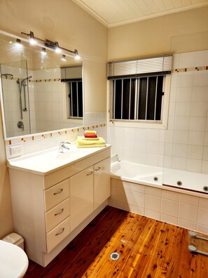 A bathroom with a wooden floor, a white sink, a mirror, a shower, and a window with blinds. - The Heights! Comfortable, cosy Queenslander cottage with a retro vibe. (Wavell Heights)