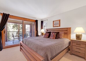 A bedroom with a large bed, a wooden headboard, a bedside table with a lamp, and a sliding glass door leading to an outdoor area with chairs.