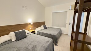 A hotel room with two beds, a nightstand, a wall-mounted lamp, and a window with a blind. - Juquehy Studios - 9 (São Sebastião)